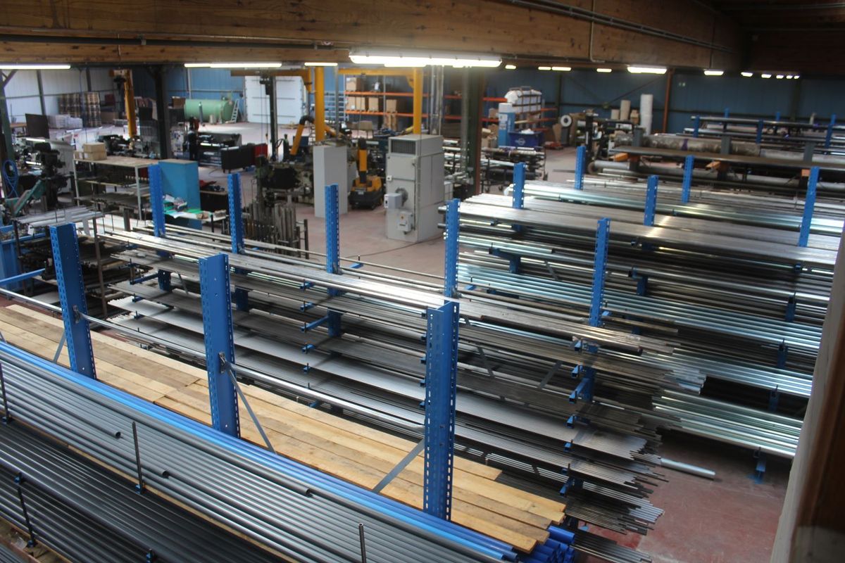 Productiehal met hoge rekken vol stalen buizen en assen, en een werkplaats met machines op de achtergrond